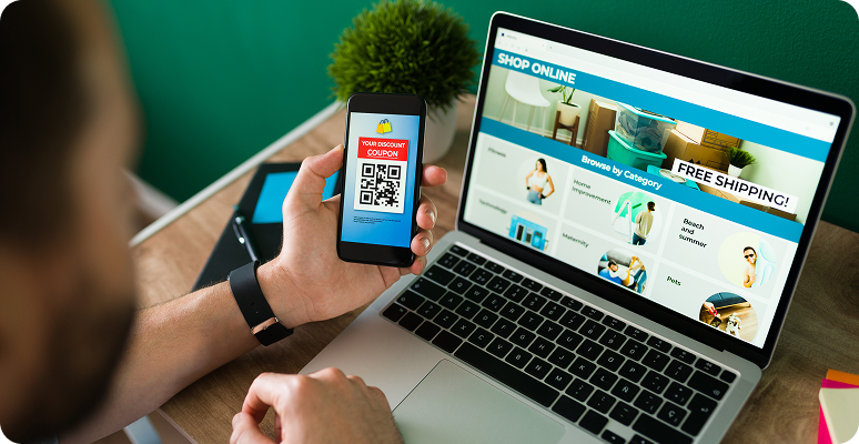 Person holding phone with QR code in front of laptop showing an online store website